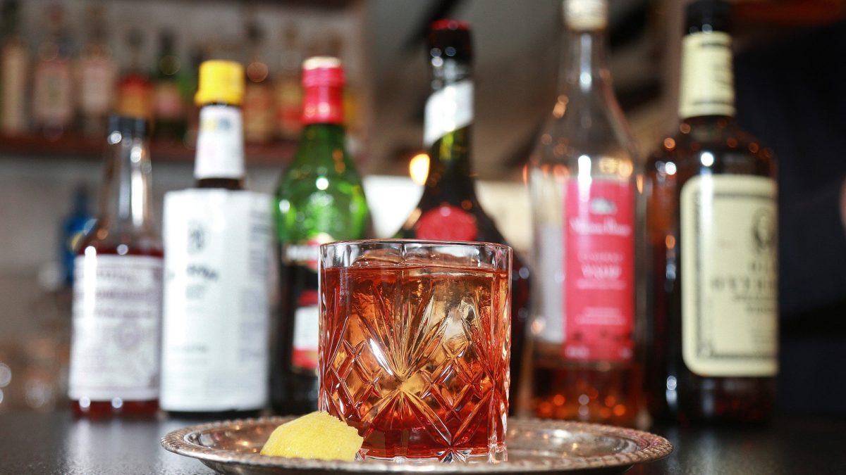 Vieux Carré Cocktail im Glas auf Metallplatte. Im Hintergrund die verwendeten Spirituosen im Glas. Daneben Zitronenscheibe. Unscharf eine Bar im Hintergrund. Frontalansicht.