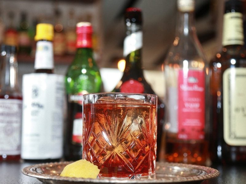 Vieux Carré Cocktail im Glas auf Metallplatte. Im Hintergrund die verwendeten Spirituosen im Glas. Daneben Zitronenscheibe. Unscharf eine Bar im Hintergrund. Frontalansicht.