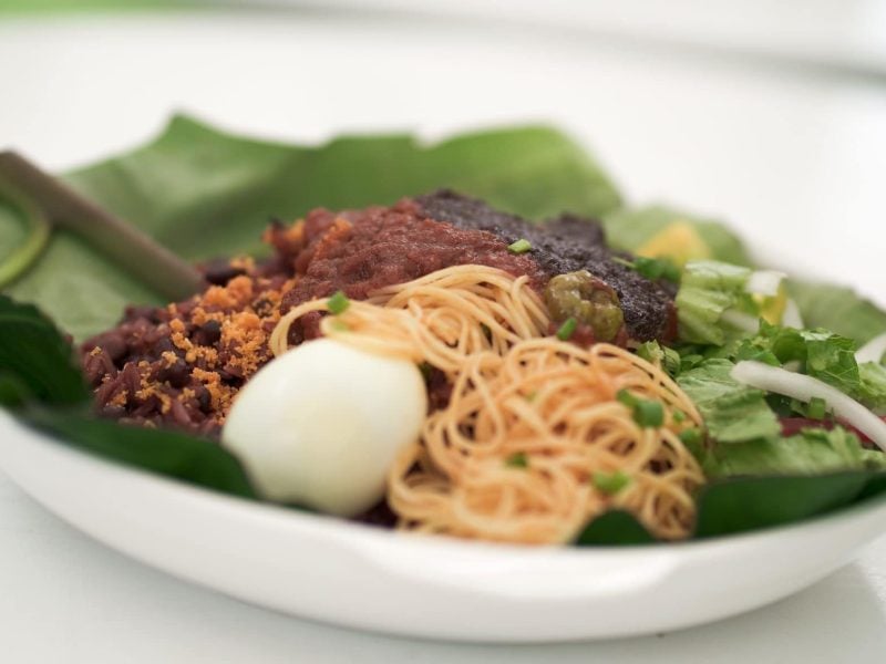 Waakye Streetfood aus Ghana mit Ghana Shito, Lammgulasch, Ei und Nudeln auf Bananenblatt auf weißem Teller. Frontalansicht.