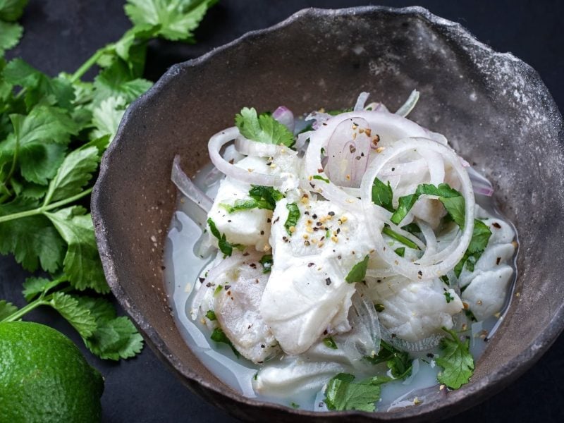 Wolfsbarsch-Ceviche auf einem grauen Teller und dunklem Untergrund. Neben dem teller ein Bund Koriander und eine Limette.