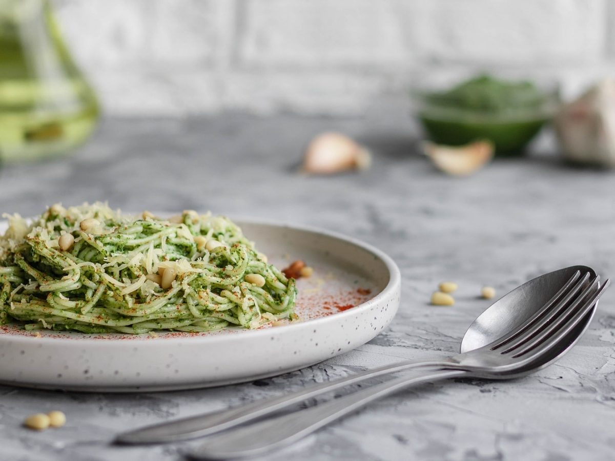 Ein Teller Zucchini-Avocado-Pasta mit Pinienkernen, daneben liegt Besteck und Knoblauch.