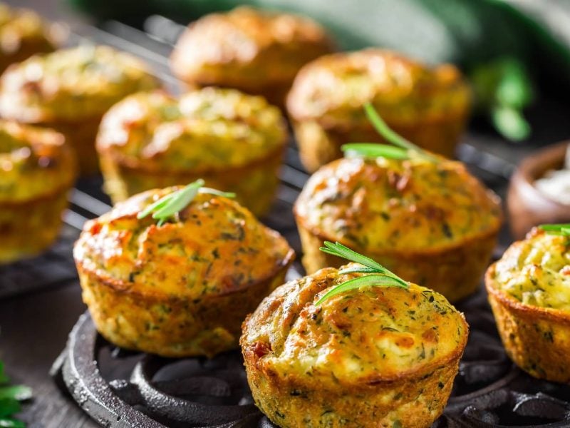 Mehrere der Zucchini-Muffins auf einem Blech mit Kräutern dekoriert.