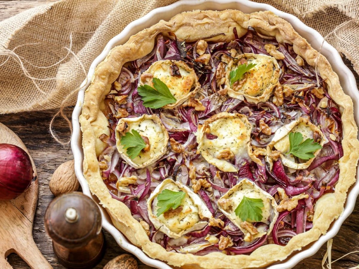 Der Zwiebelkuchen auf einem rustikalem Holztisch. Daneben liegend strohfarbener Leinenstoff, Petersilie und rote Zwiebeln.