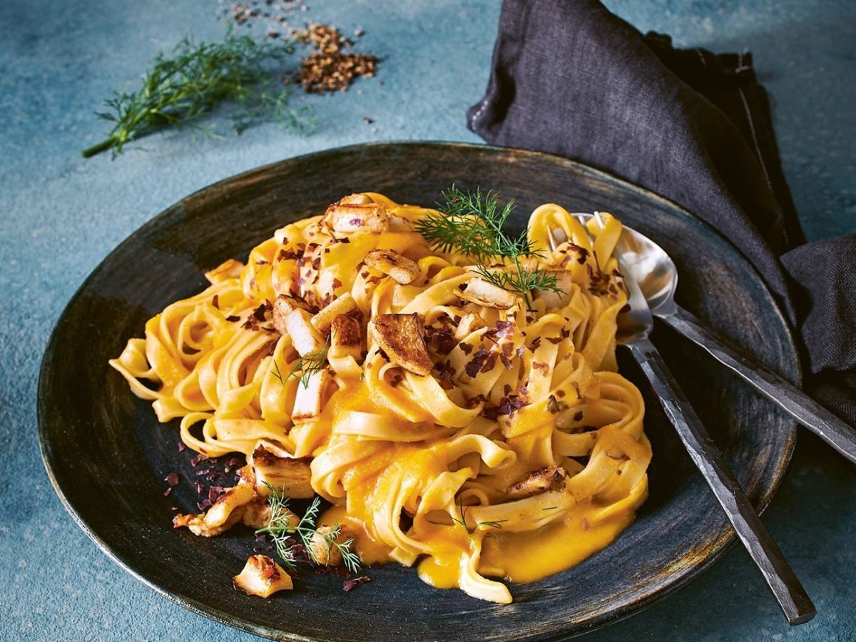 Rötliche Tagliatelle auf einem schwarzen Teller und blauem Untergrund.