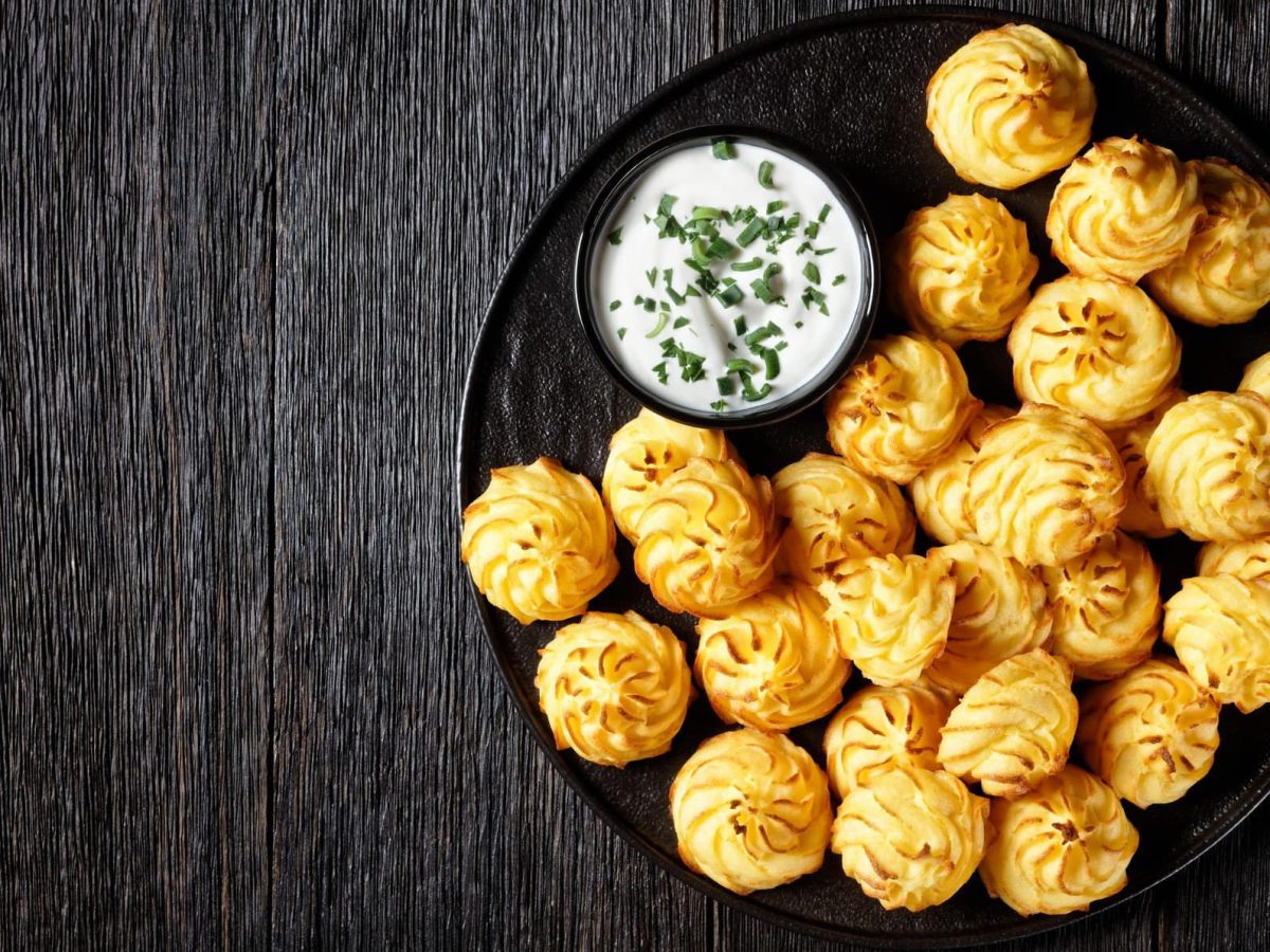 Ein schwarzer Teller mit Herzoginkartoffeln neben einem Dip auf dunklem Hintergrund.