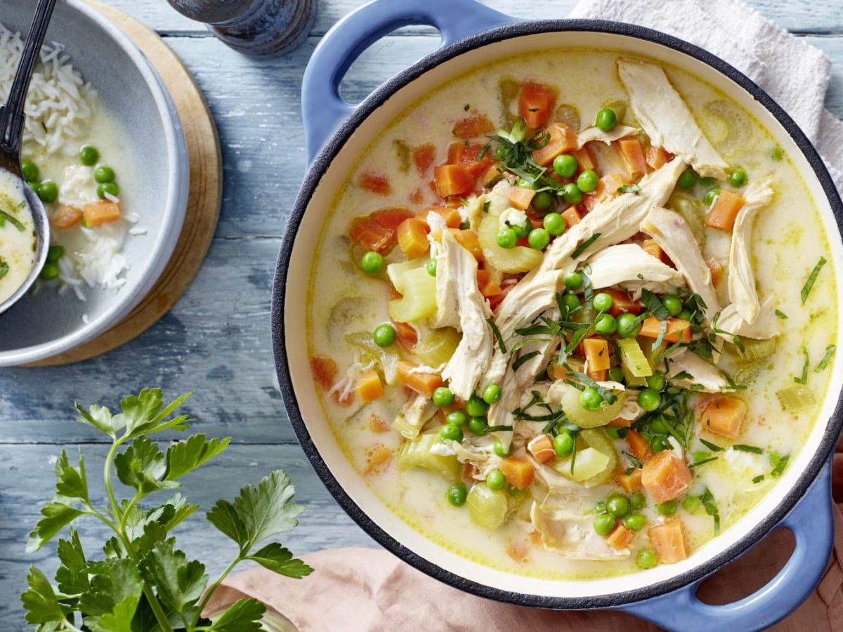 Griechische Hühnersuppe mit Petersilie im Topf neben einer Schüssel mit Reis und einer Kelle Suppe vor blauem Hintergrund,