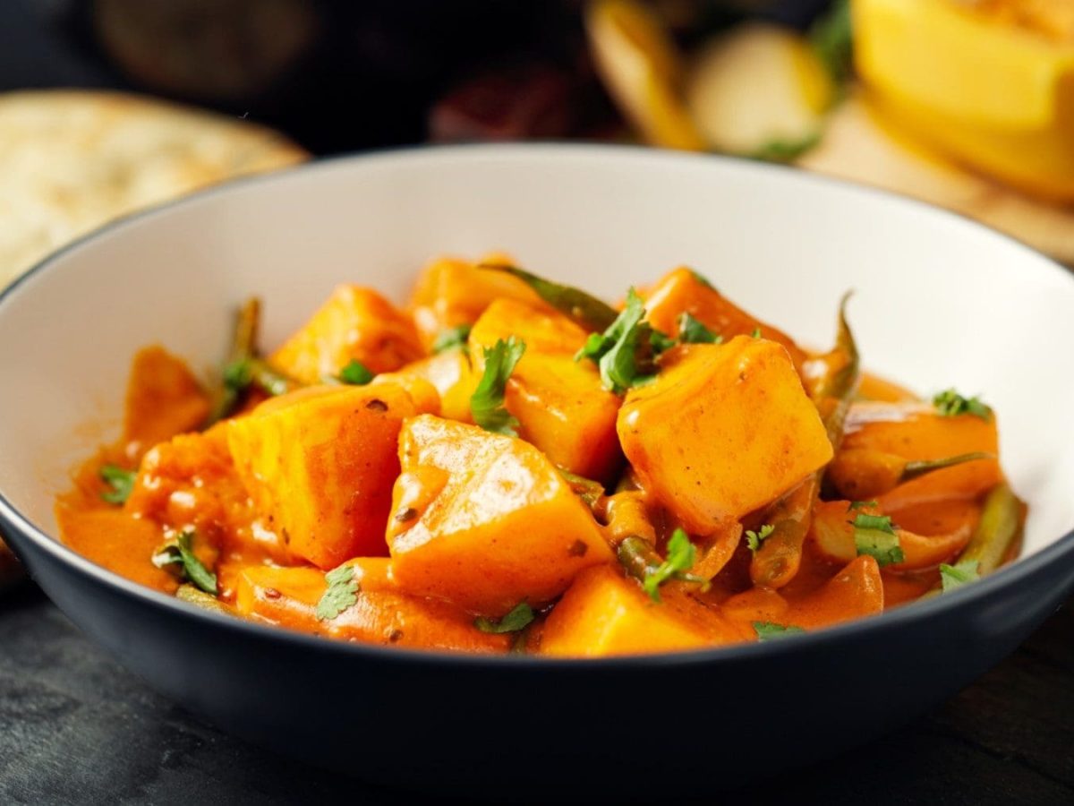 Tiefer Teller mit Kartoffel-Curry mit Kürbis auf schwarzem Untergrund, im Hintergrund ist Brot Kürbis zu sehen.