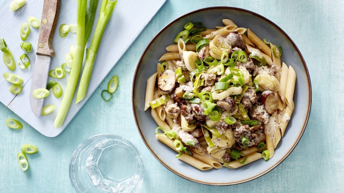 Pasta mit Pilzen, Lauch und Hack, garniert mit frischen Lauchzwiebeln und serviert in einer hellen Schale auf blauem Tisch.