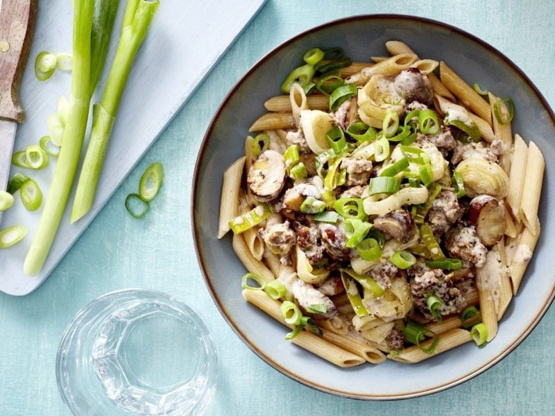 Pasta mit Pilzen, Lauch und Hack, garniert mit frischen Lauchzwiebeln und serviert in einer hellen Schale auf blauem Tisch.