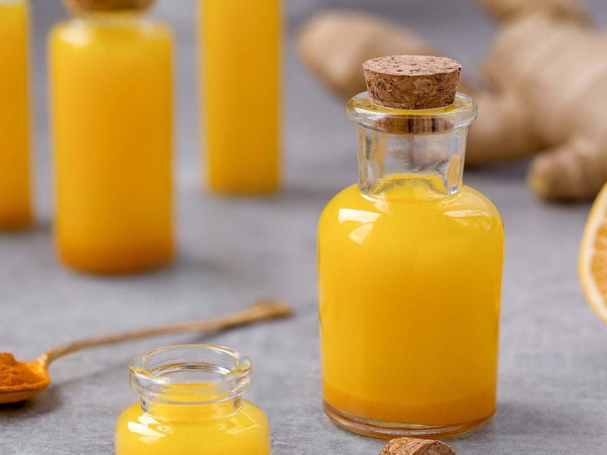 Mehrere Flaschen Ananas-Ingwer-Shots, drumherum ein Löffel Kurkuma, Ingwerwurzeln und eine Orange.