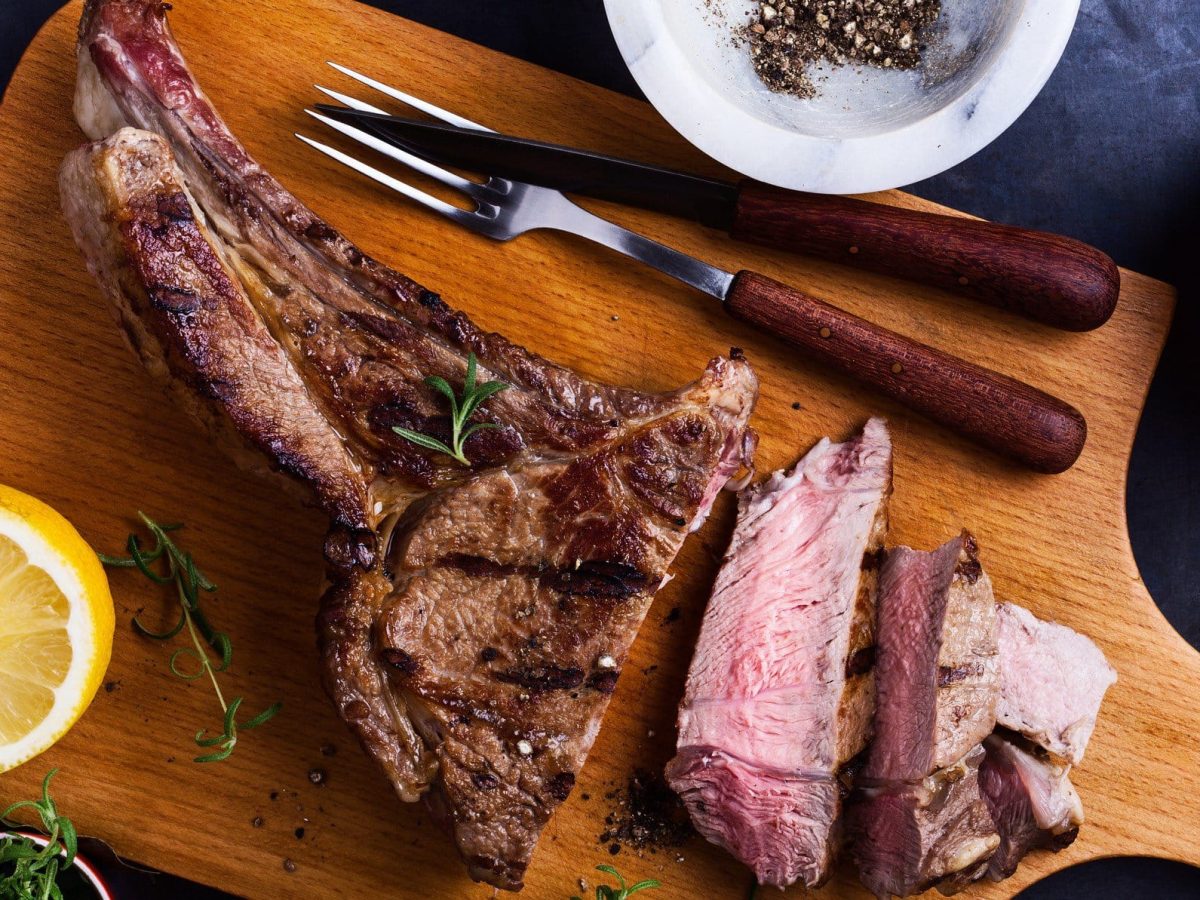 Anleitung zum Rückwährtsgaren für Entrecote Double am Knochen. Aufgeschnittenes Steak auf Holzbrett mit Besteck. Daneben eine Zitrone, Kräuter und je eine Schale mit schwarzem Pfeffer und grobem Salz. Draufsicht.