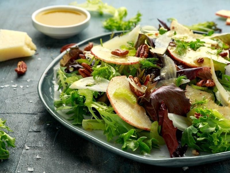 Frontalansicht: EIn Teller Apfel-Fenchel-Salat steht auf einem festen Untergrund. Drumherum liegen ein Stück Käse, eine Schale mit einer Flüssigkeit und Salatblätter.