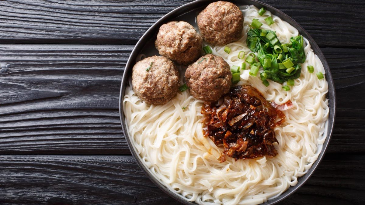 Indonesische Nudelsuppe mit Hackbällchen Bakso in Rinderbrühe mit Sambal-Zwiebeln in schwarzer Suppenschüssel auf dunklem Holz. Draufsicht-