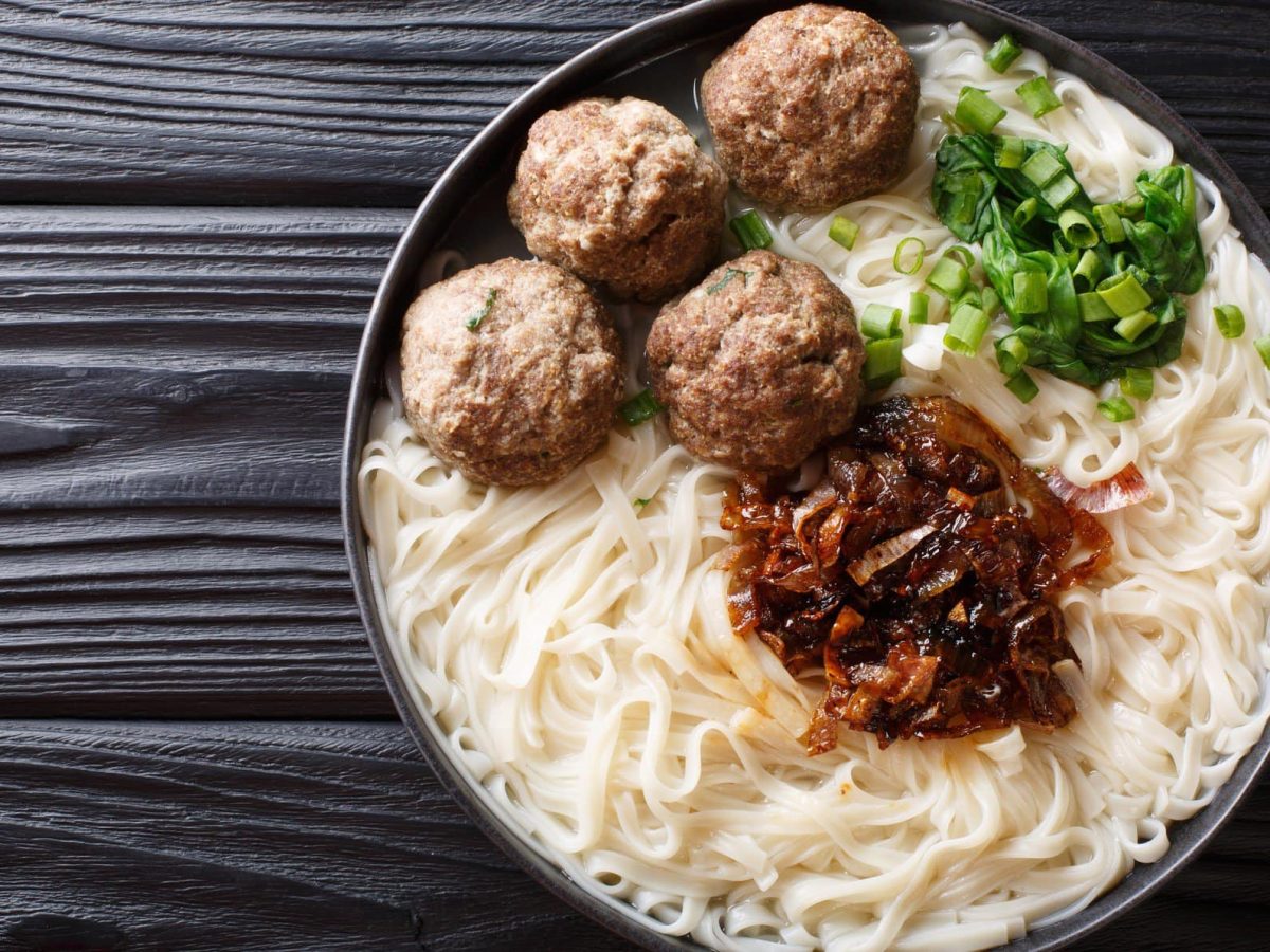 Indonesische Nudelsuppe mit Hackbällchen Bakso in Rinderbrühe mit Sambal-Zwiebeln in schwarzer Suppenschüssel auf dunklem Holz. Draufsicht-