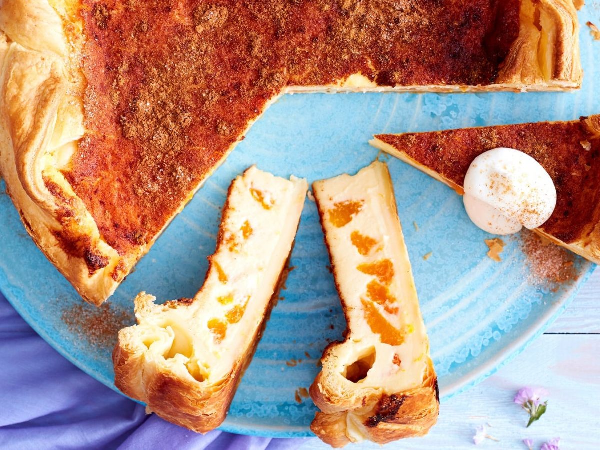 Drei Stücke vom Blätterteig-Schmand-Kuchen mit dem restlichen Kuchen auf einem blauen Teller vor blauem Hintergrund.