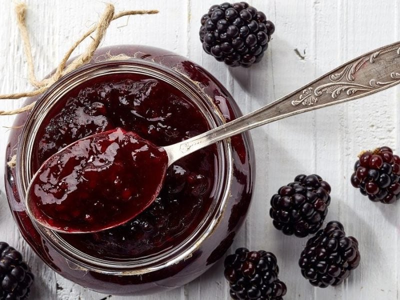 Vogelperspektive: In einem Glas Brombeer-Gelee mit Vanille steckt ein Löffel. Drumherum liegen auf der weißen Fläche einige frische Brombeeren.