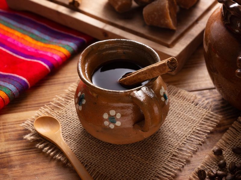 Café de Olla traditioneller mexikanischer Kaffe in Tongefäß mit Zimtstange auf Stofftuch. Daneben traditionelles mexikanisches Tischtuch, brauner Zucker, eine Tonkanne und Zimtstangen. Draufsicht.