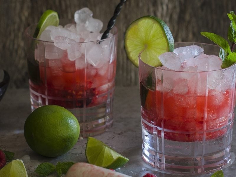 Zwei Gläser Caipiroska mit Himbeeren mit Minz garniert, neben Limetten vor grauem Hintergrund.