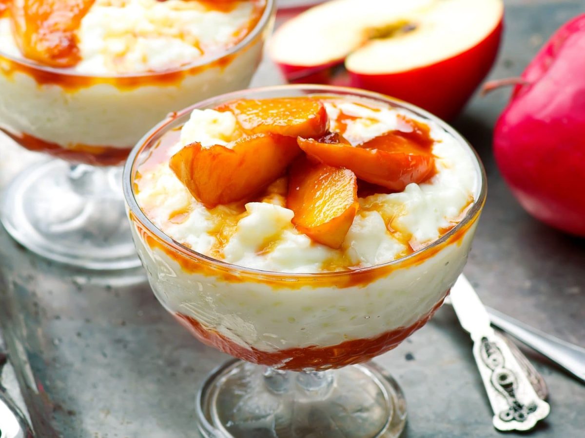 Zwei Dessertgläser Chai-Milchreis mit Zimtäpfeln, daneben liegt ein halbierter Apfel.