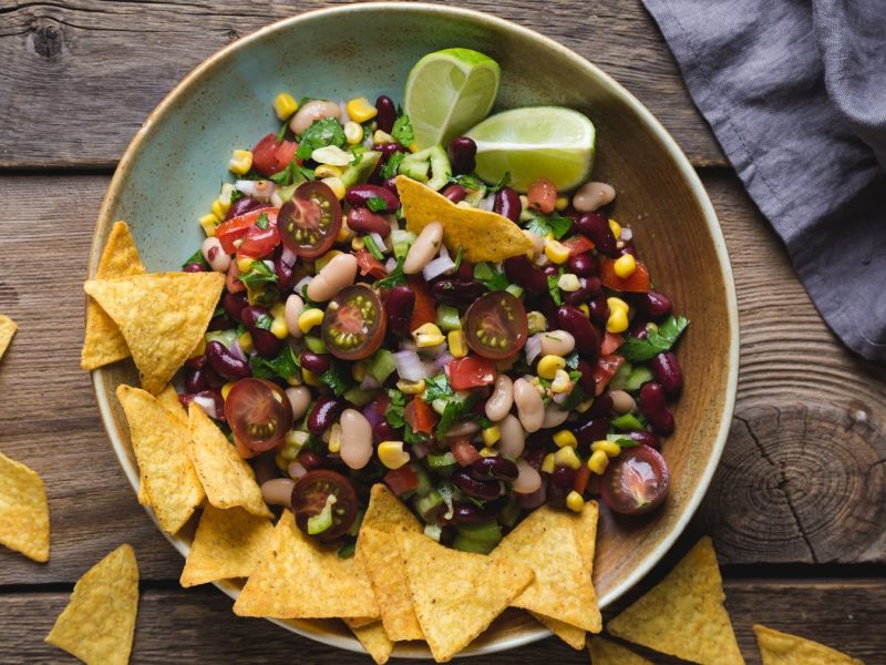 Eine Schale Cowboy Caviar, ein Dip oder Salat aus Bohnen, Tomaten, Mais, Zwiebeln und Paprika, dazu Nachos und Limettenspalten.