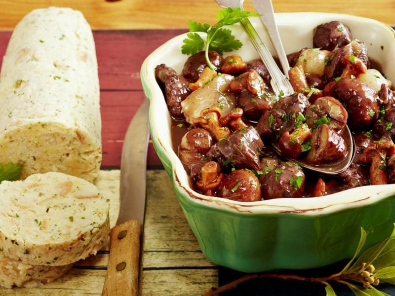 Damwild-Gulasch mit Maronen und Waldpilzen in grüner Auflaufform auf Holzbrett. Daneben Serviettenknödel und Petersilie. Draufsicht.