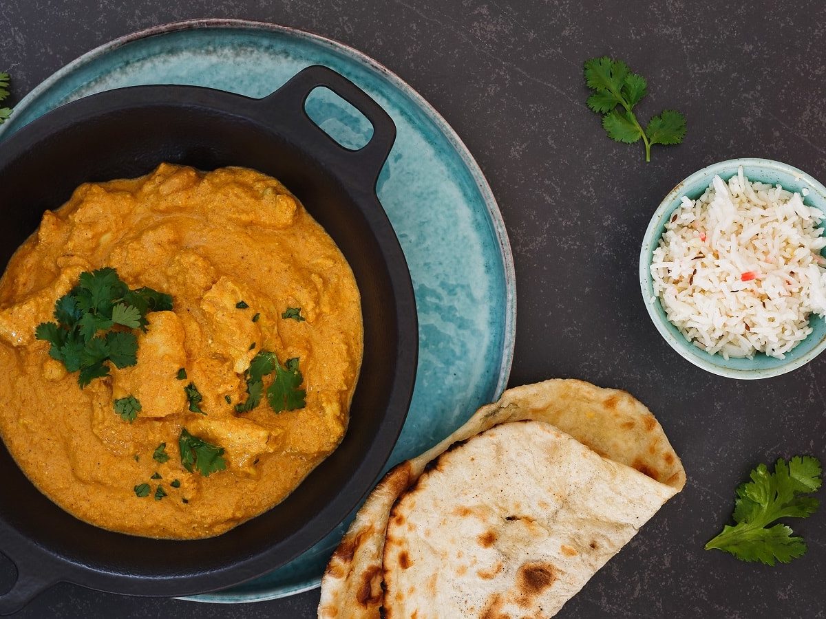 Eine schwarze Schüssel mit Chicken Korma auf einem hellblauen Teller neben Naan, Reis und frischem Koriander.