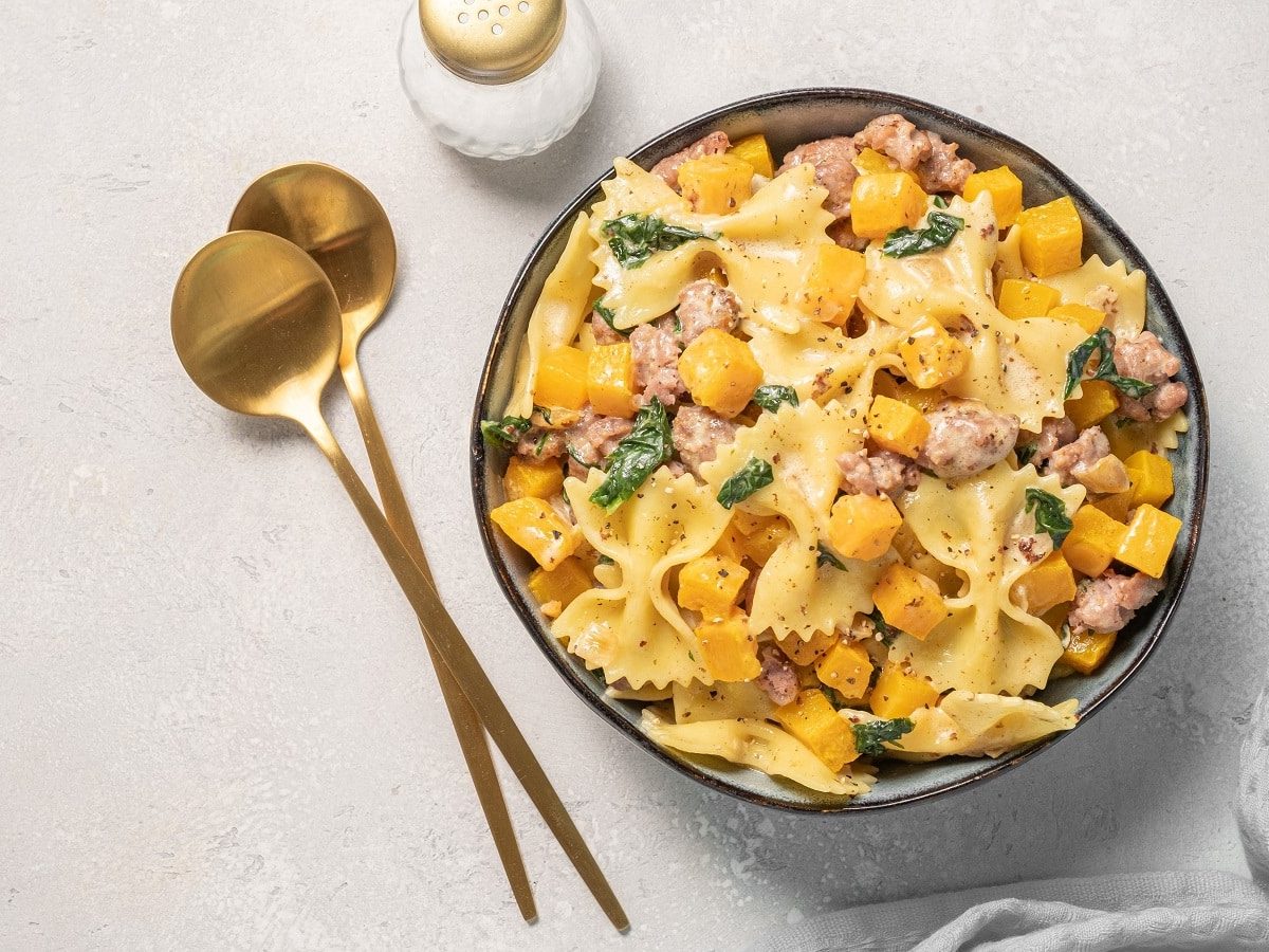 Ein runder Teller mit Farfalle mit Kürbis neben roségoldenem Besteck.