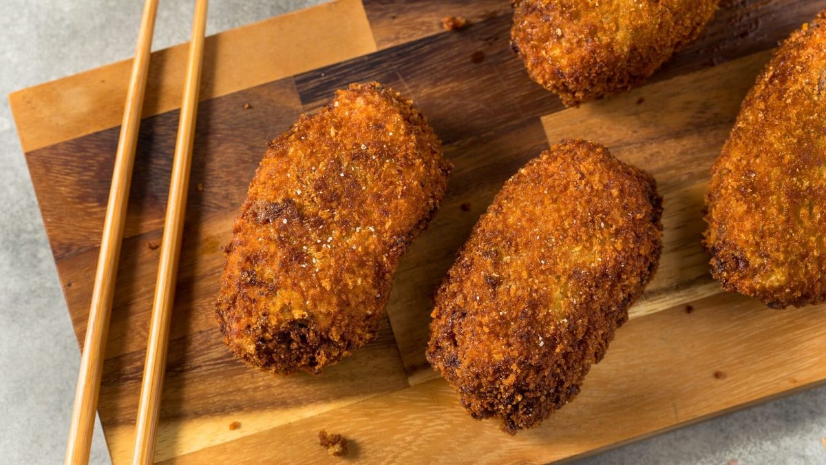 Ein Holzbrett mit Korokke darauf neben einem Paar Stäbchen.