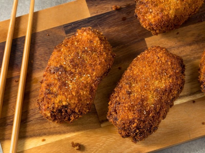 Ein Holzbrett mit Korokke darauf neben einem Paar Stäbchen.