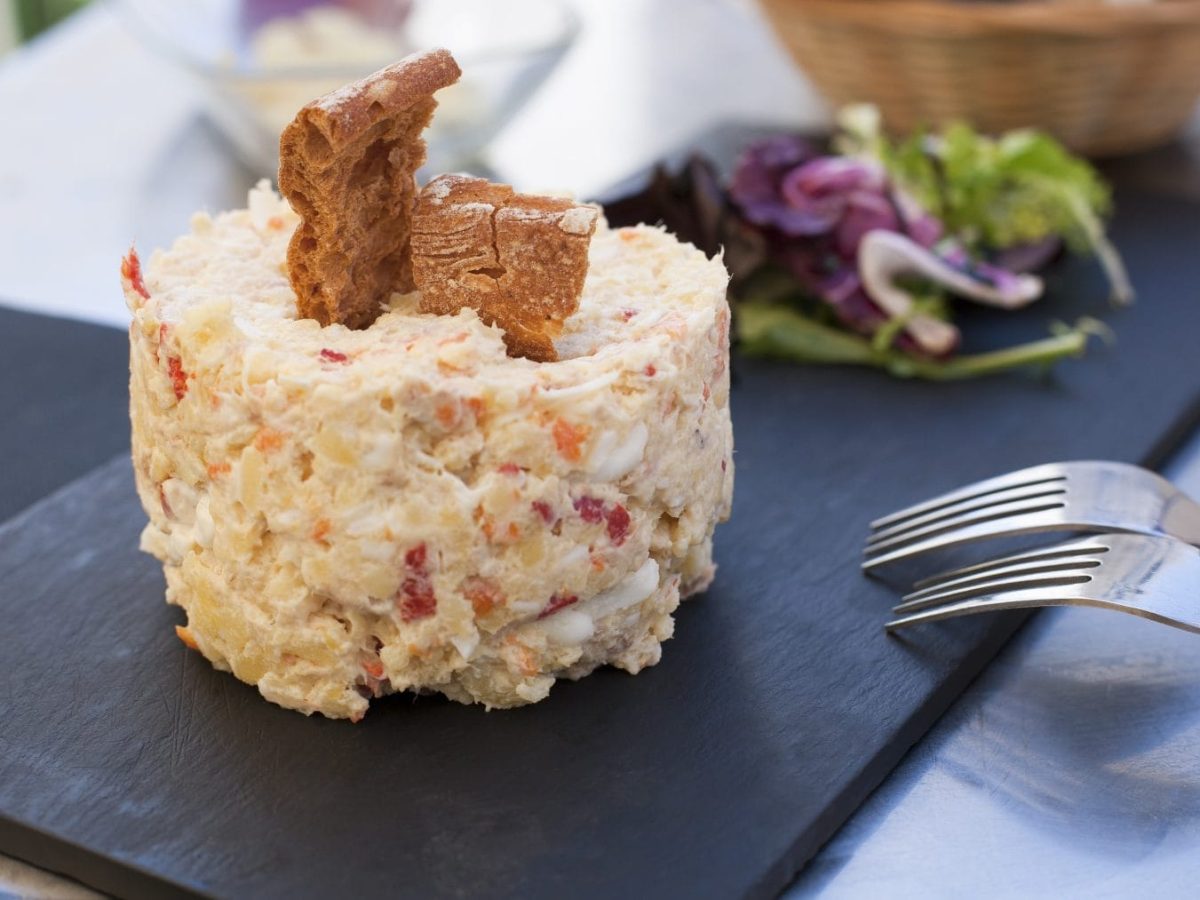 Ensalada Rusa angerichtet mit Salat und Brotchips auf Schieferplatte. Daneben zwei Gabeln. Frontalansicht.