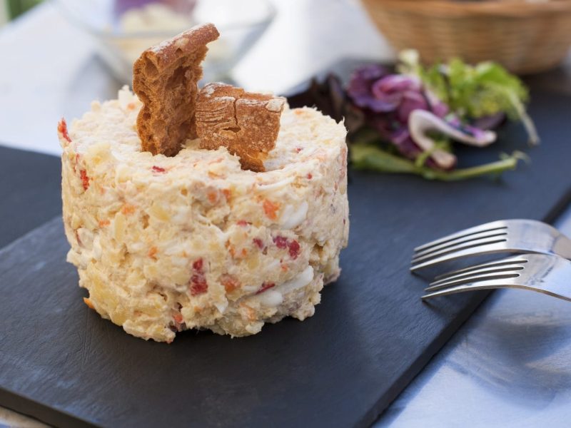 Ensalada Rusa angerichtet mit Salat und Brotchips auf Schieferplatte. Daneben zwei Gabeln. Frontalansicht.