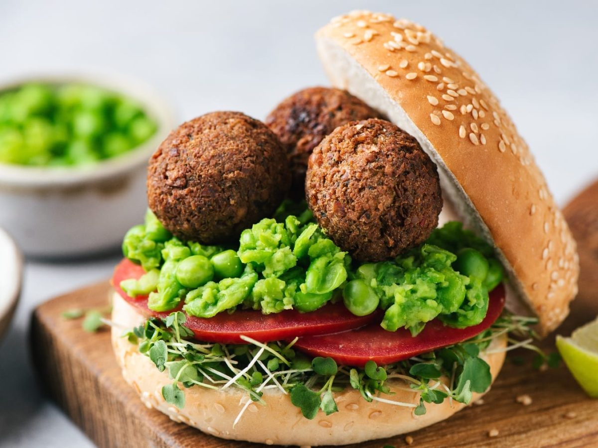 Ein aufgeklapptes Burgerbrötchen mit drei Falafelkugeln und etwas Erbsen-Guacamole auf einem kleinen Holzschneidebrett..