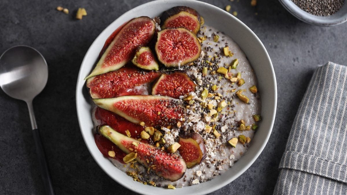 Eine weiße Schale mit dem Granola und frischen Feigen auf einem dunkelgrauen Untergrund. Links daneben ein Metalllöffel udn rehcts daneben eine graue Serviette.