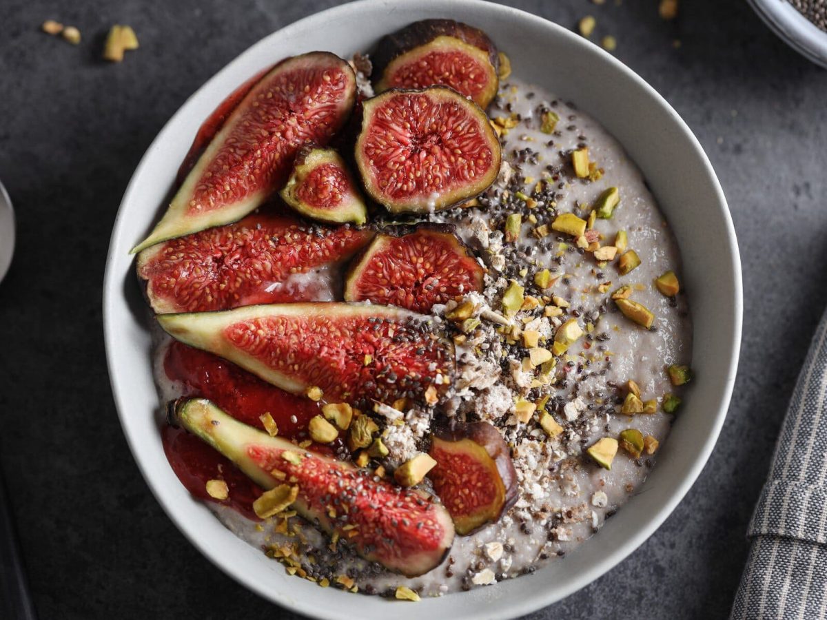 Eine weiße Schale mit dem Granola und frischen Feigen auf einem dunkelgrauen Untergrund. Links daneben ein Metalllöffel udn rehcts daneben eine graue Serviette.