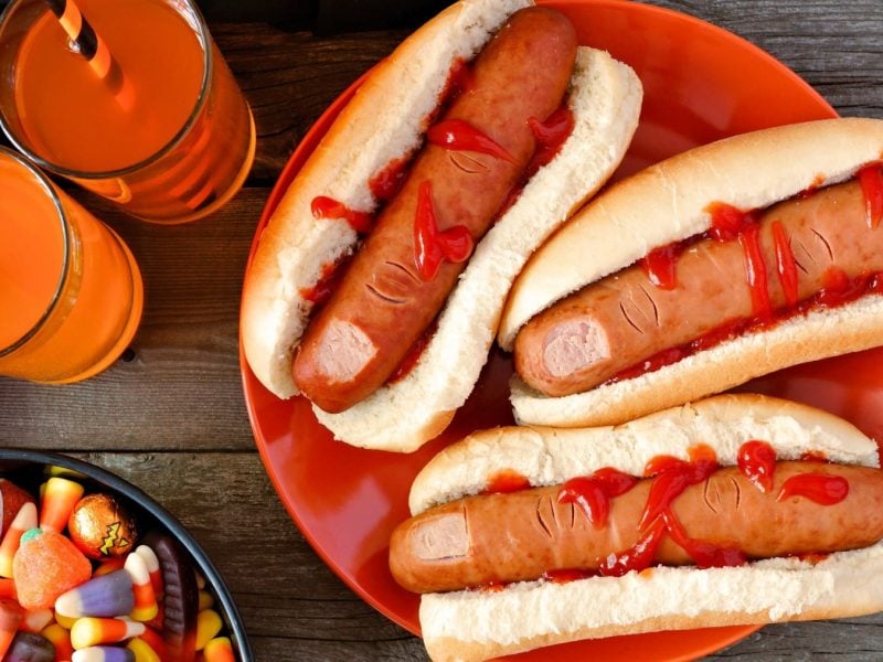 Drei Finger Hot Dogs mit Ketchup auf einem orangenen Teller, neben orangenen Getränken und Süßigkeiten vor grauem Holzhintergrund.