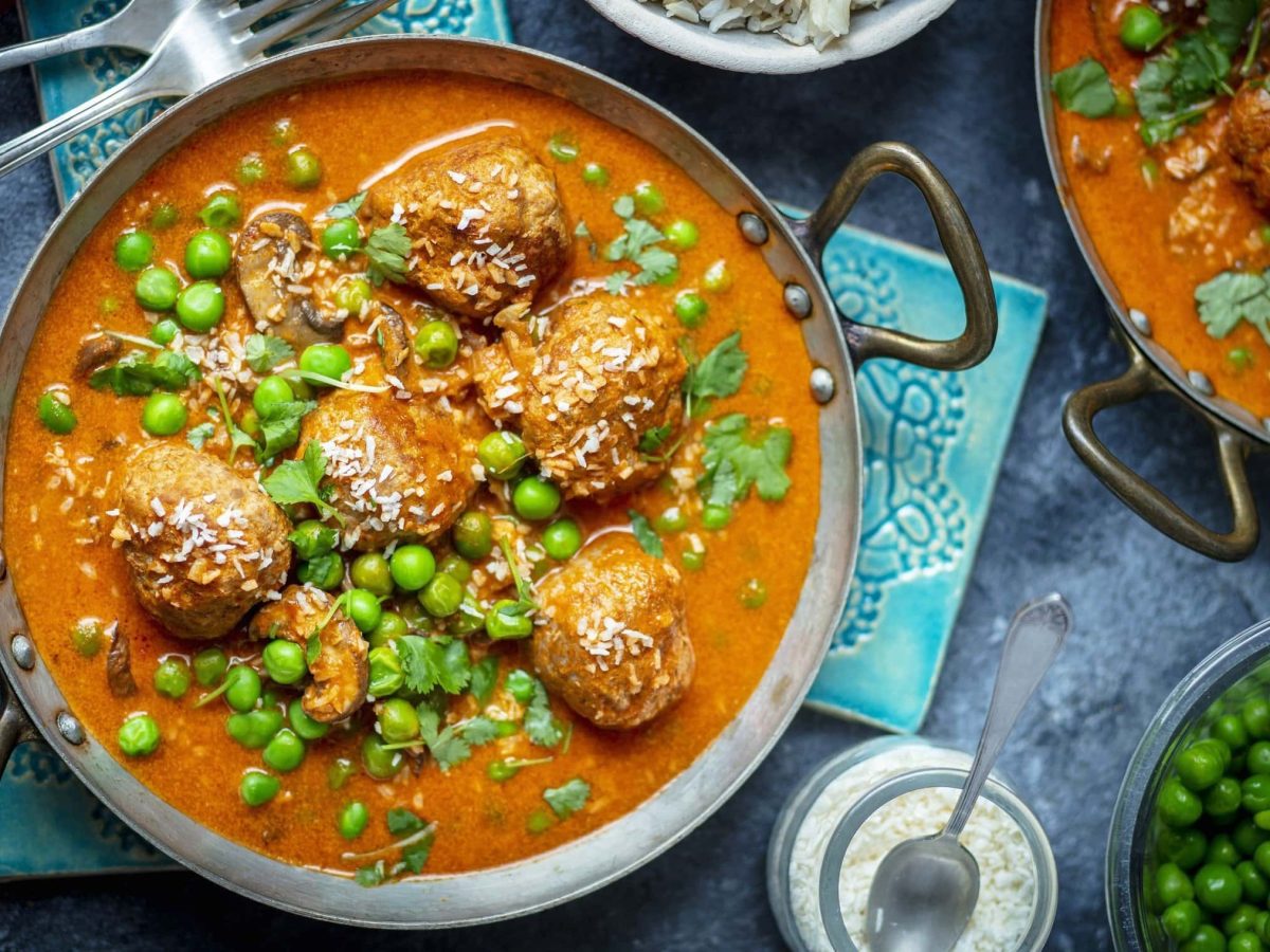 Fleischbällchen in Currysauce mit grünen Erbsen in einem Topf neben Zutaten vor blauem Hintergrund.