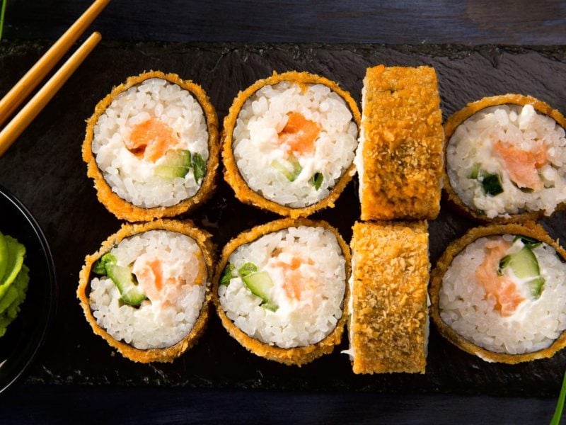 Acht Fried Sushi Röllchen vor dunklem Hintergrund neben Wasabi und Stäbchen.