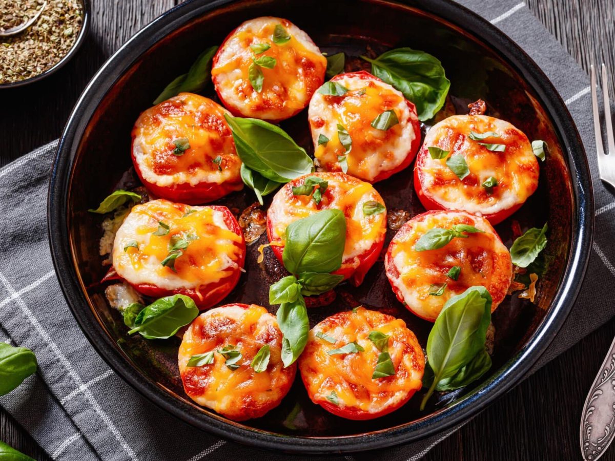 Gebackene Tomaten mit dreierlei Käse auf schwarzem Teller mit Basilikum auf grauem Tuch und Tisch. Oben Schale mit Oregano. Daneben Besteck und Basilikum. Draufsicht.