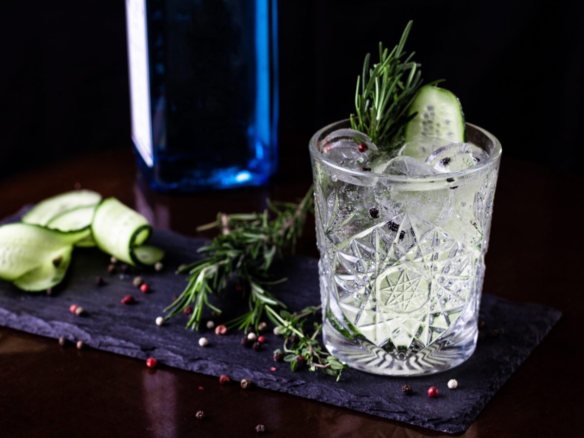 Zum Gin Tonic Day Gin Tonic im Glas mit Rosmarin und Gurke auf Schieferplatte auf Holz. Daneben Gin Flasche, Pfeffer und Gurkenscheiben. Frontalansicht.