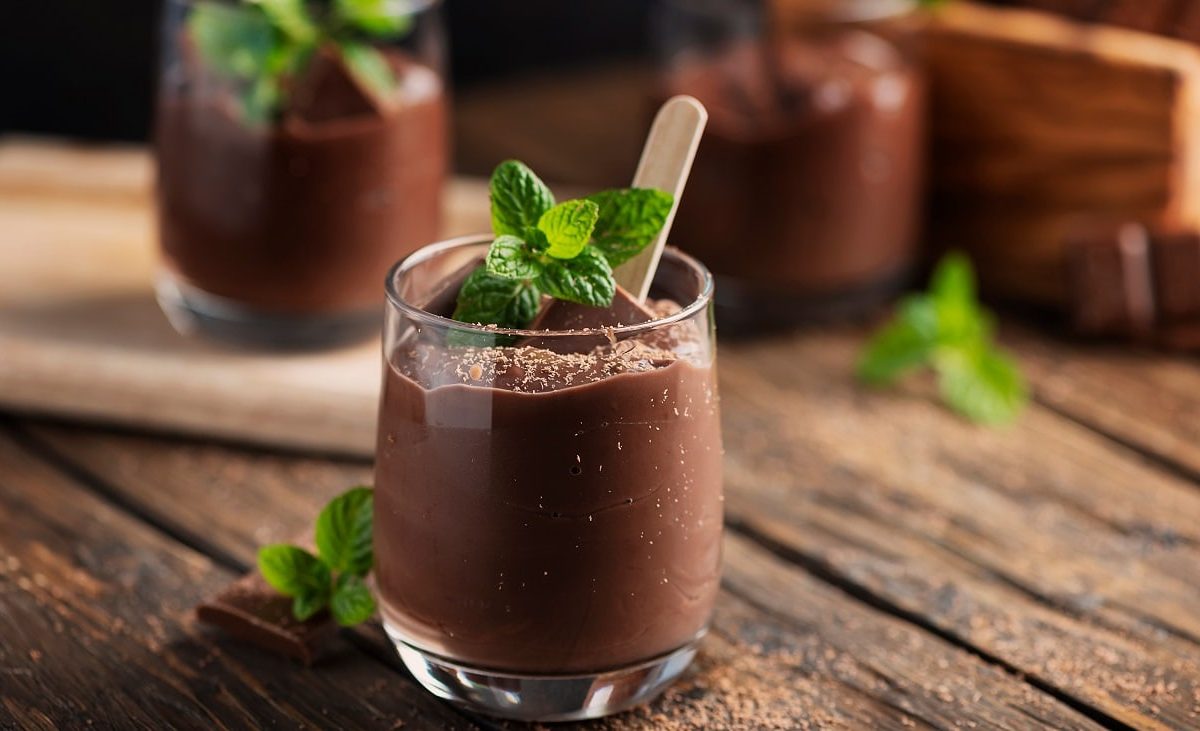 Frontalsicht: Zwei Gläser mit einem dunklen Pudding stehen auf einem Untergrund aus Holz. Auf dem Pudding ist ein Blatt Minze.