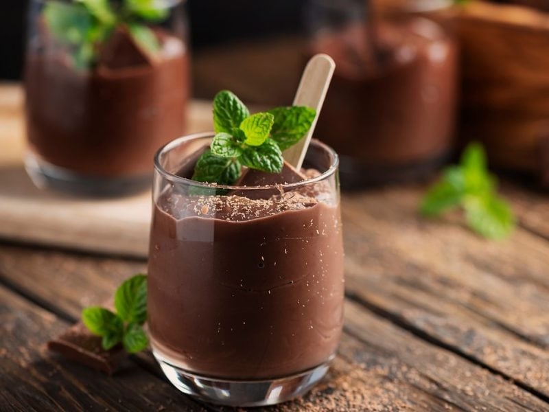 Frontalsicht: Zwei Gläser mit einem dunklen Pudding stehen auf einem Untergrund aus Holz. Auf dem Pudding ist ein Blatt Minze.