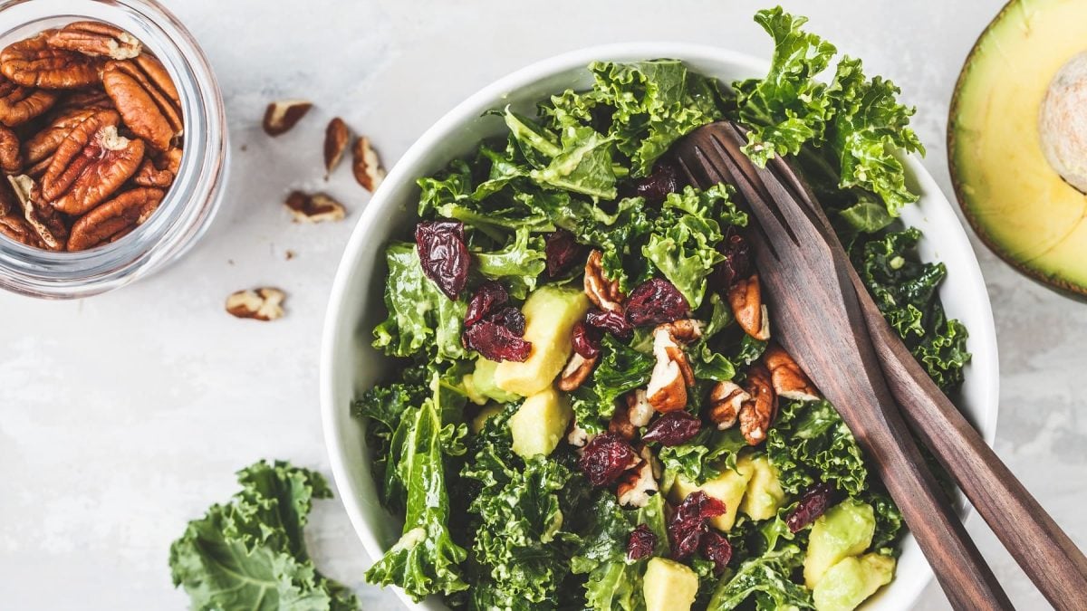 Grünkohlsalat mit Avocado, Birne, Pecannüssen und Cranberrys in der Draufsicht, darin Salatbesteck, daneben Nüsse und eine halbe Avocado.