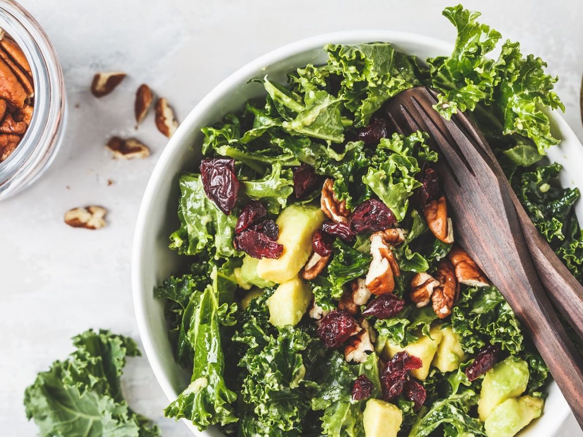 Grünkohlsalat mit Avocado, Birne, Pecannüssen und Cranberrys in der Draufsicht, darin Salatbesteck, daneben Nüsse und eine halbe Avocado.