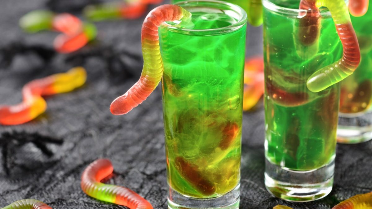 Shotgläser mit grünen Halloween Jello Shots mit Gummiwürmern auf schwarzer Fläche mit mehr Würmern im Hintergrund.