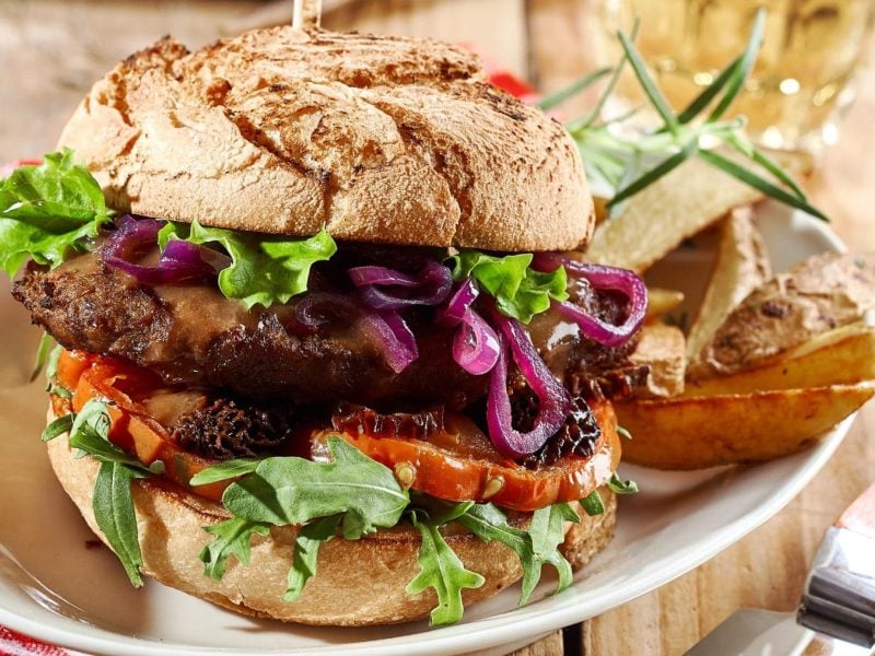 Wildburger vom Hirsch im Malzbrötchen auf Teller mit Kartoffelecken. Im Hintergrund ein Bierglas, rot-weiße Stoffserviette. Vorne Messer mit Holzgriff. Frontalansicht.