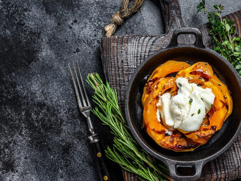 Gebackene Hokkaidospalten mit Burrata in schwarzer Auflaufform auf grauem Tuch. Daneben Kräuter und eine Schale mit pinkem Salz. Draufsicht.