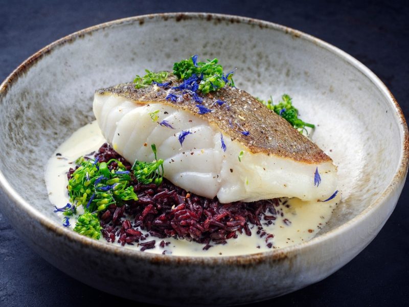 Ein grauer Teller mit schwarzem Reis und darauf ein Stück Kabeljau. Als Saucenspiegel eine helle Sauce udn alles dekoriert mit etwas Brokkoli und blauen Kornblumenblüten.