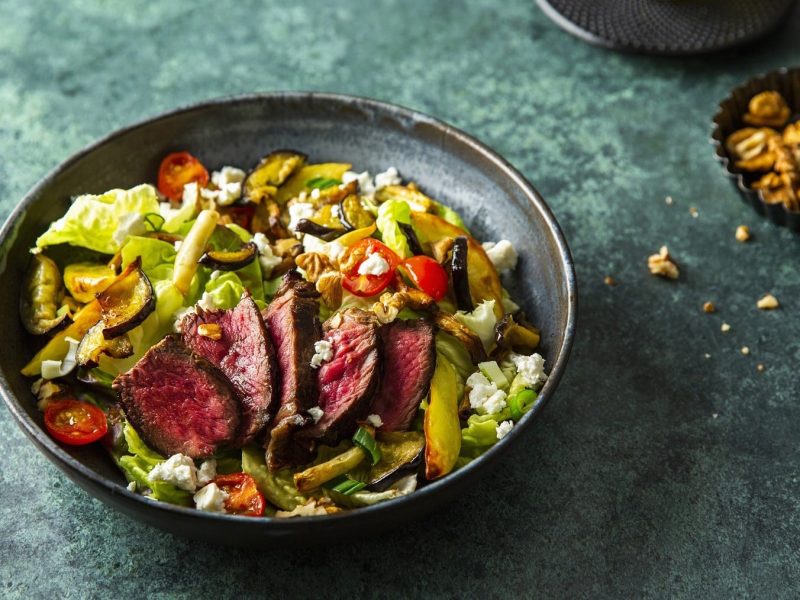 Kaffeemariniertes Steak auf Grillgemüse-Salat mit Nüssen und Feta in einem schwarzen Teller vor dunklem Hintergrund.