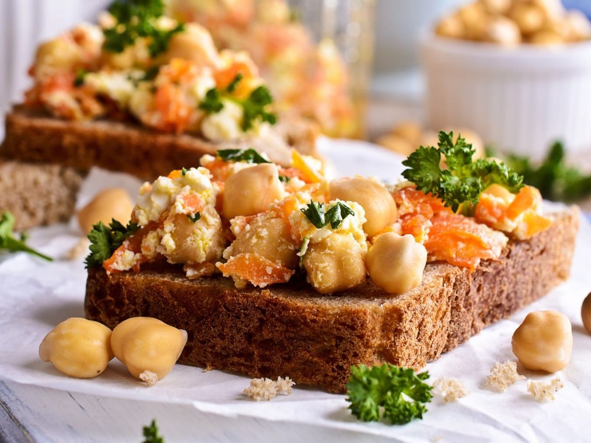 Zwei Kichererbsen-Karotten-Sandwiches auf Butterbrotpapier, im Hintergrund eine Schale Kichererbsen.