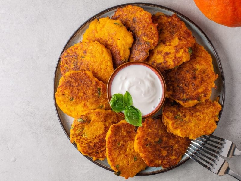 Mehrere Kürbis-Hashbrowns auf einem Teller übereinanderliegend im Kreis angeordnet und in der Mitte eine kleine Schale mit einem weißen Dip. Alles auf einem hellgrauen Untergrund.